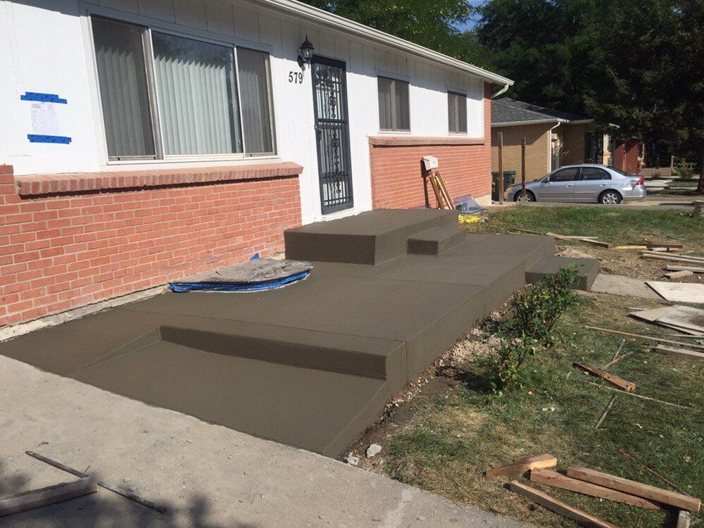 Concrete ramp leading to a front door of a brick house, with unfinished steps and a parked car nearby.
