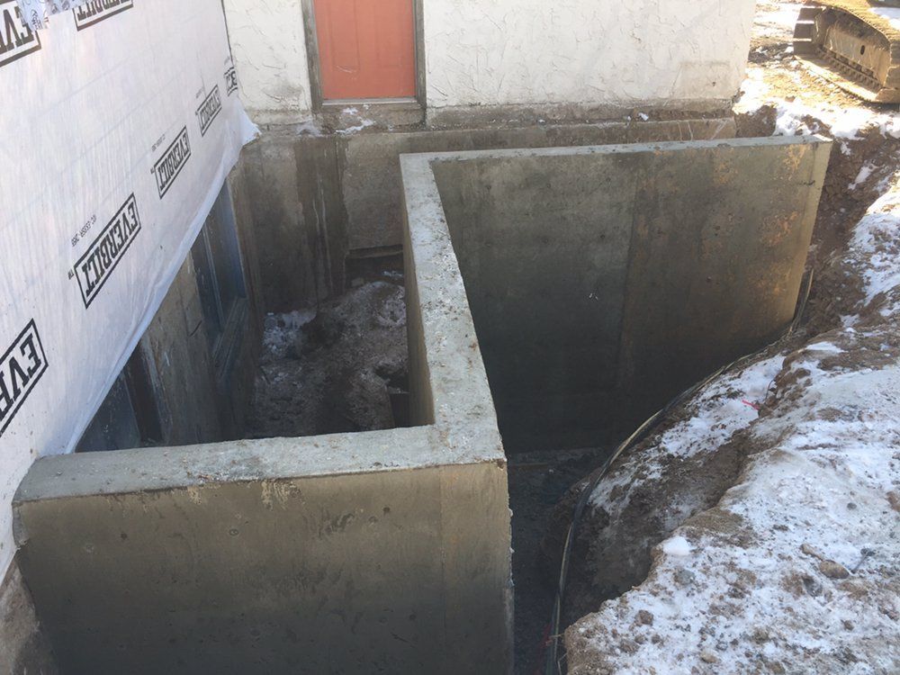 Concrete foundation walls under construction beside a house, with snow on the ground and house wrap visible.