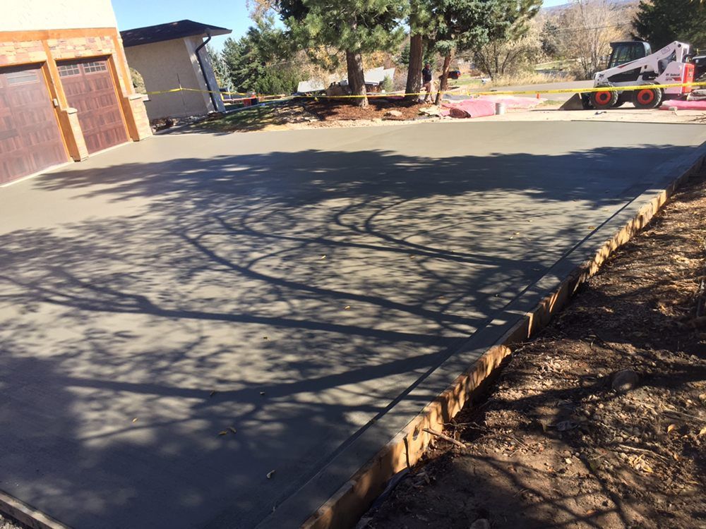 Concrete driveway in front of a house, bordered by mulch and trees in bright sunlight.
