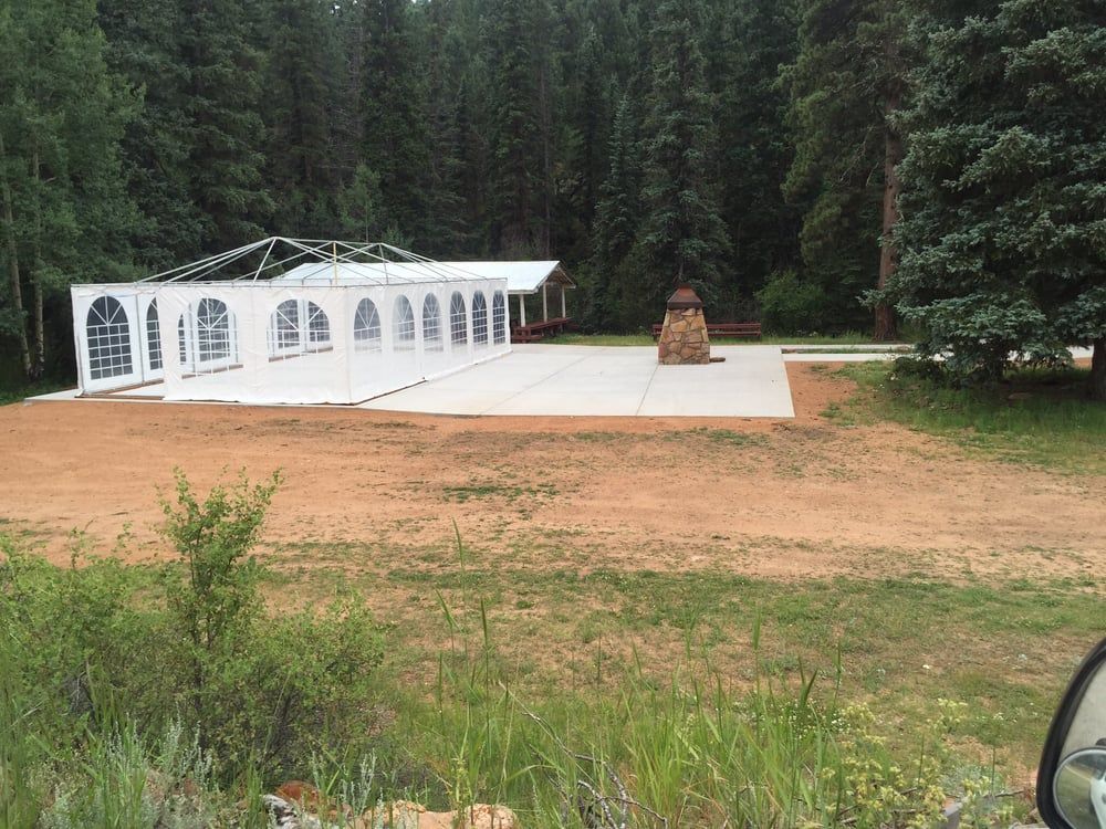 White arched tent on concrete pad in a forest clearing, with a small wooden structure nearby.