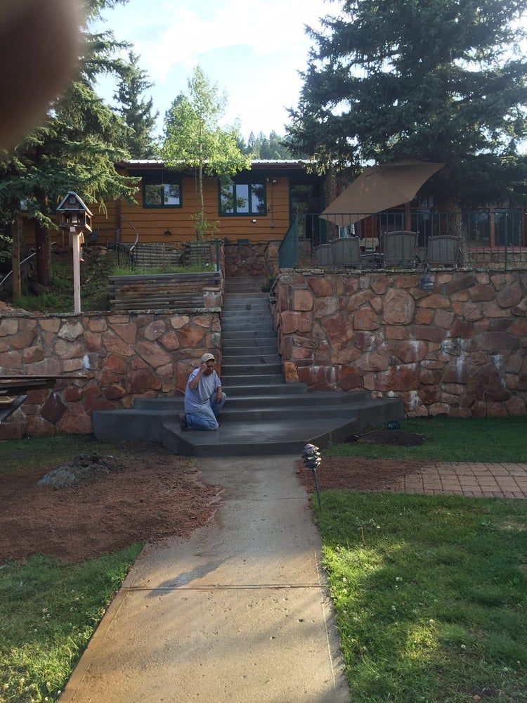 Stone steps lead to a wooden house on a hill, with a child seated on the steps and a small dog below.