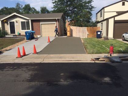 Residential driveway newly paved, with orange cones, a wooden fence, and trash bins near a house.