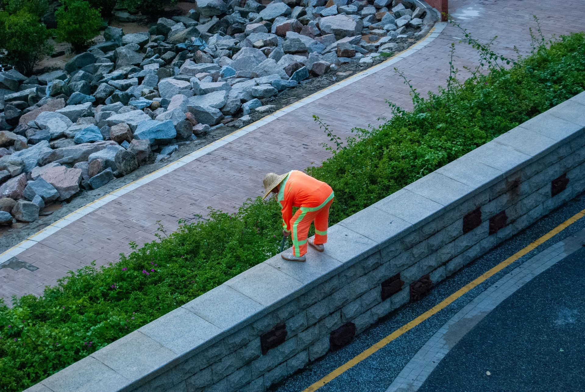 Worker in bright orange outfit tending grass on a sloped roadside beside a stone wall.