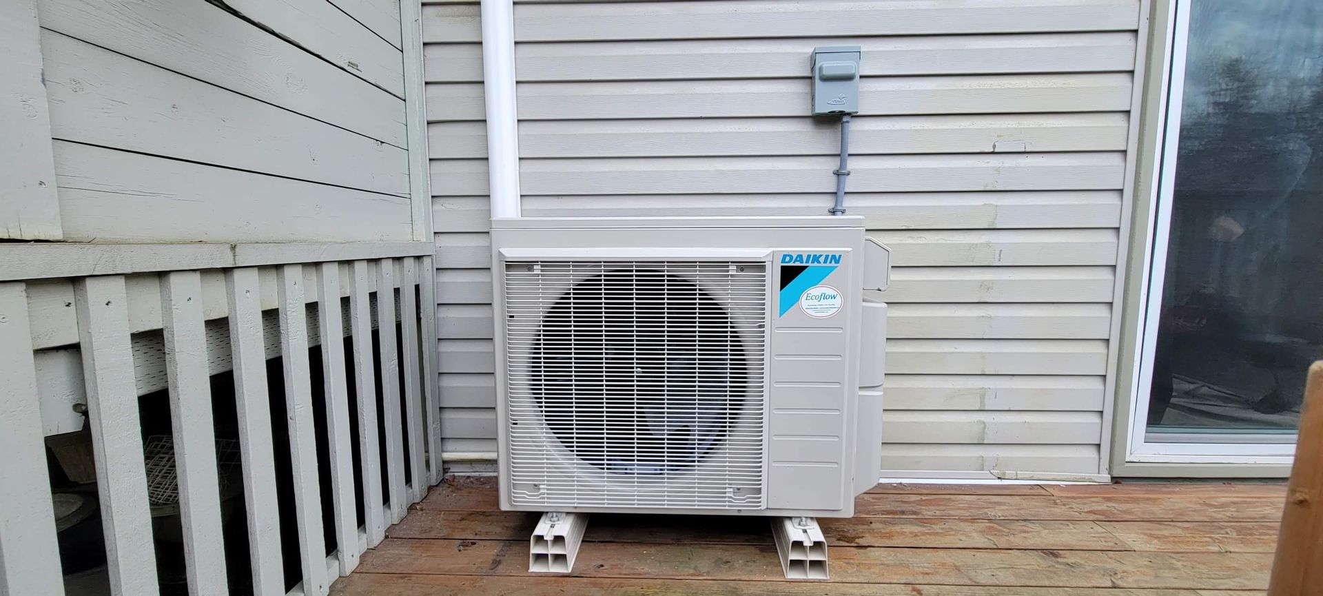 An air conditioner is sitting on a wooden deck next to a house.