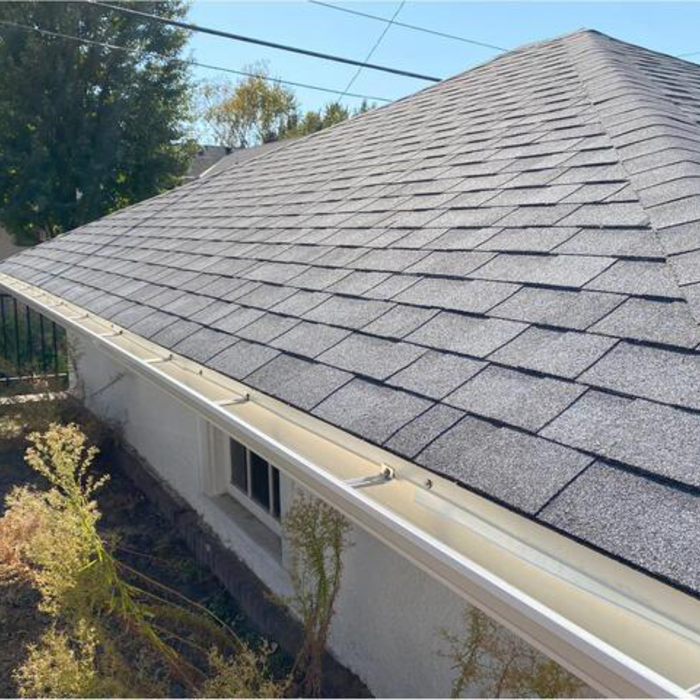 Gray asphalt shingle roof with white gutters on a light-colored building under a blue sky.