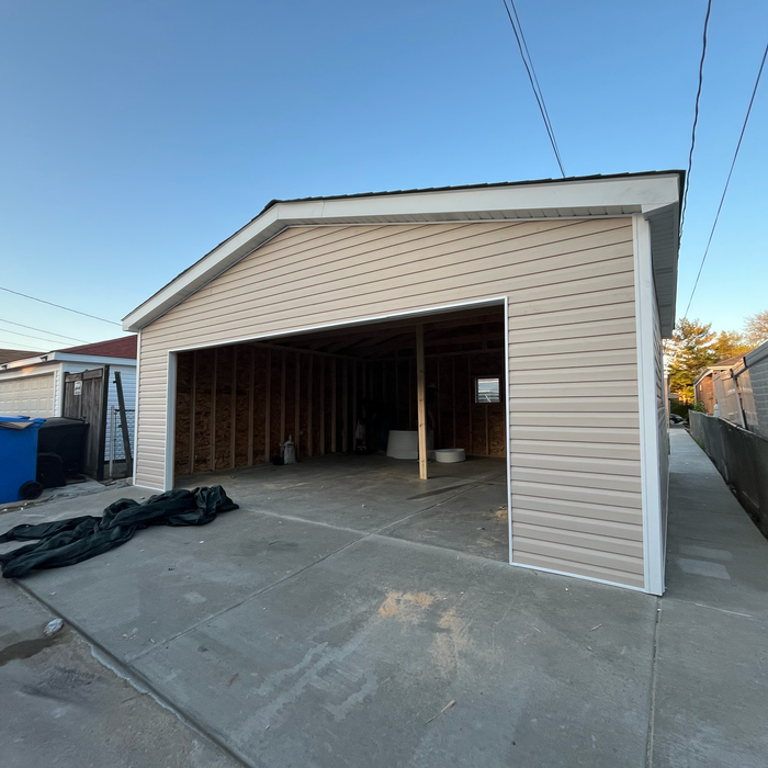 Tan-sided unfinished garage with open door, concrete floor, wooden frame, and white trim against a blue sky.