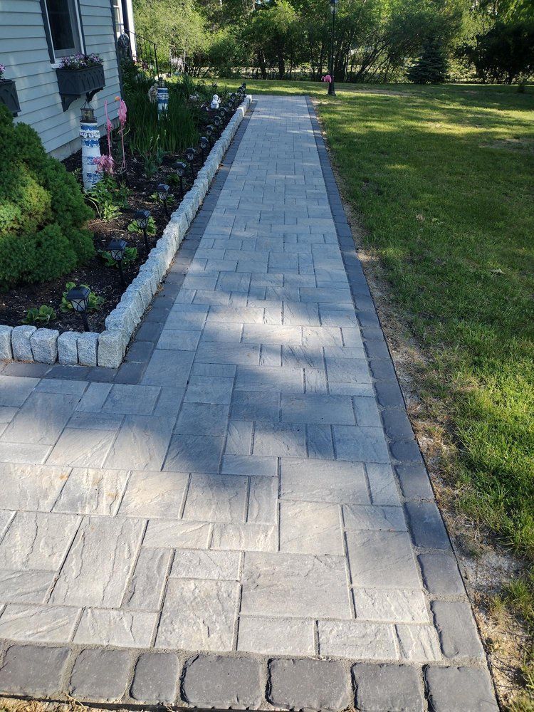 Paved stone walkway beside a house, bordered by garden beds and lawn in bright sunlight.
