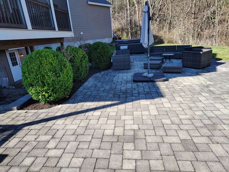 Stone patio with round fire pit, umbrella, and neatly trimmed green shrubs beside a house