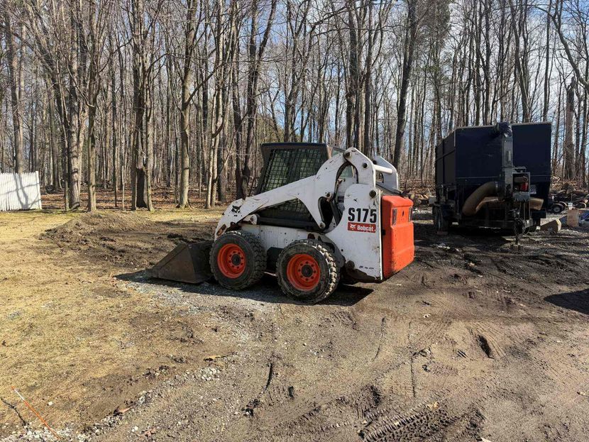 Skid steer loader with orange wheels at a wooded worksite, with another machine and truck nearby.