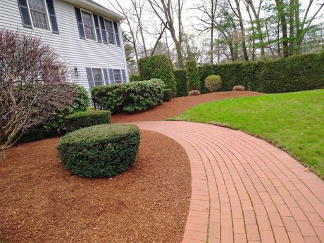 Curved brick path through mulched garden beds beside a white house and green lawn