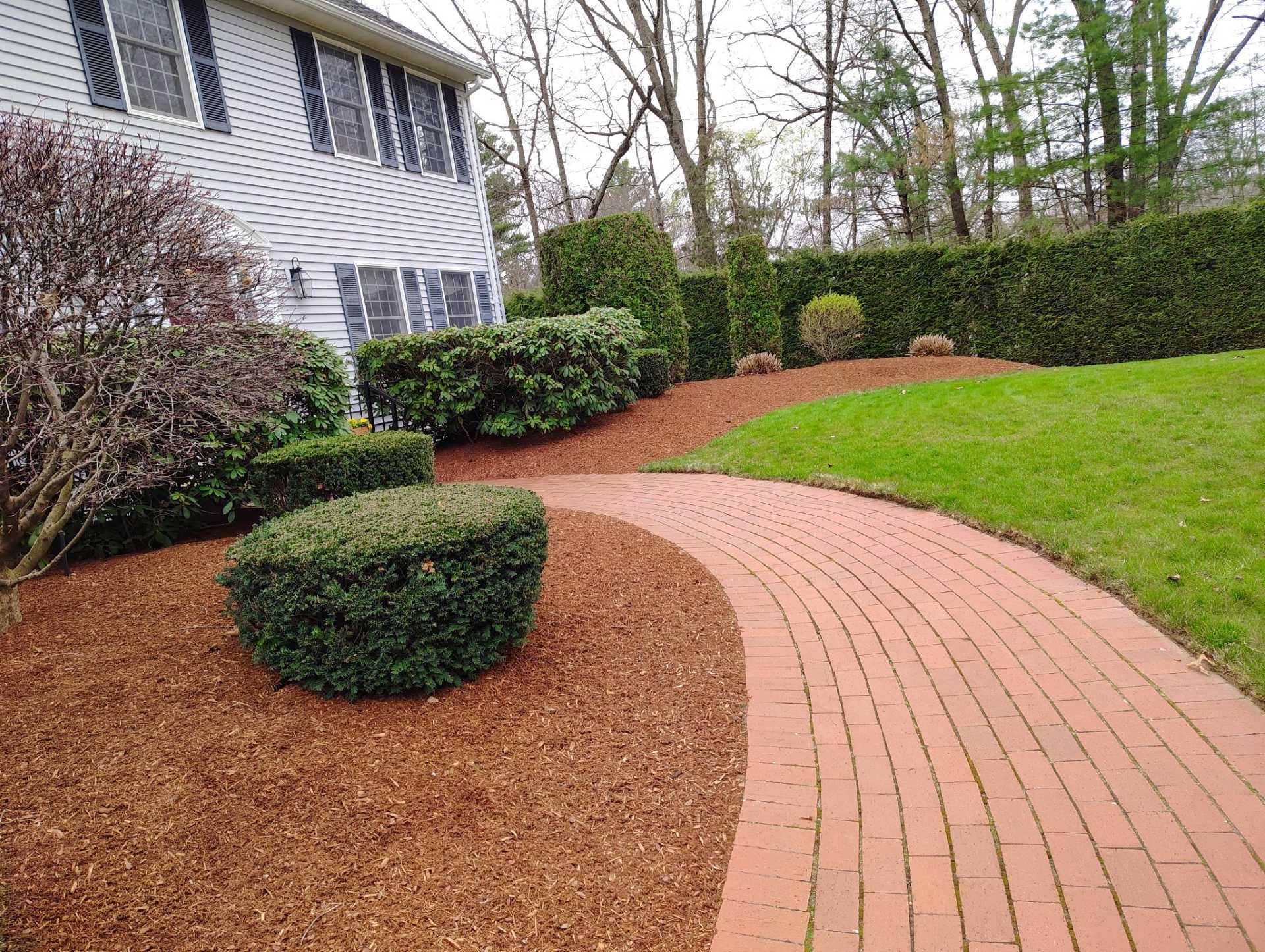 Curved brick walkway beside a house, lined with mulch, shrubs, and green lawn.