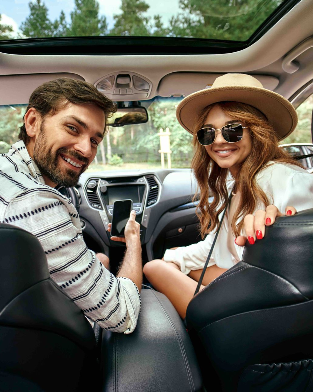 A couple smiling inside a car with a sunroof, the man holding a phone, both looking at the camera.