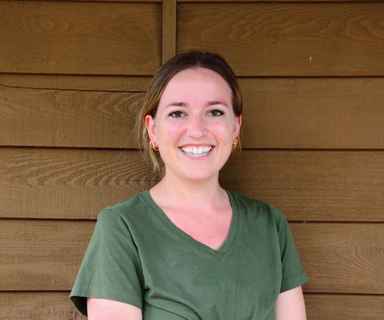 Woman with a wide smile wearing a green v-neck shirt, in front of a brown wooden wall.