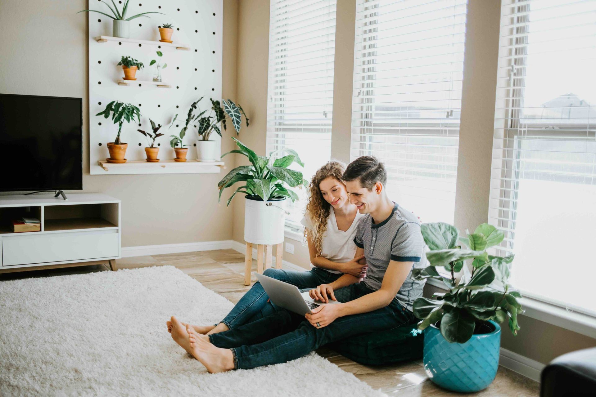 Couple sitting on a rug, looking at laptop. Plants and a large window in a bright living room.
