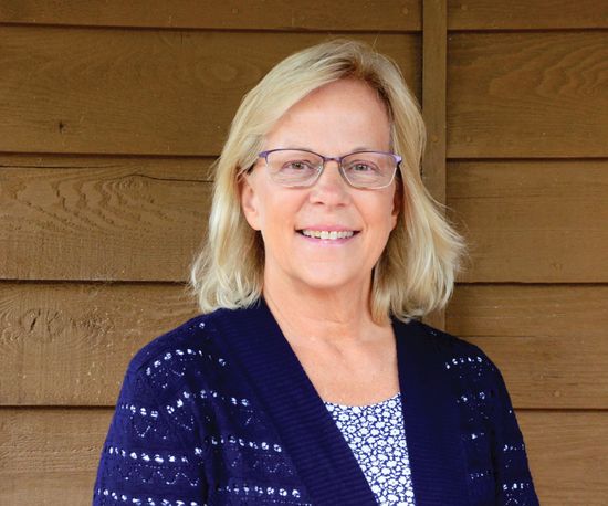 Woman with glasses smiles, wearing blue cardigan and patterned top, standing in front of wood siding.