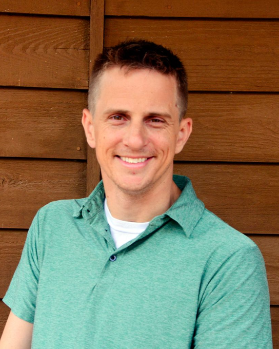 Eric Chase, smiling, wearing a green polo shirt and a white undershirt, in front of a wood background.