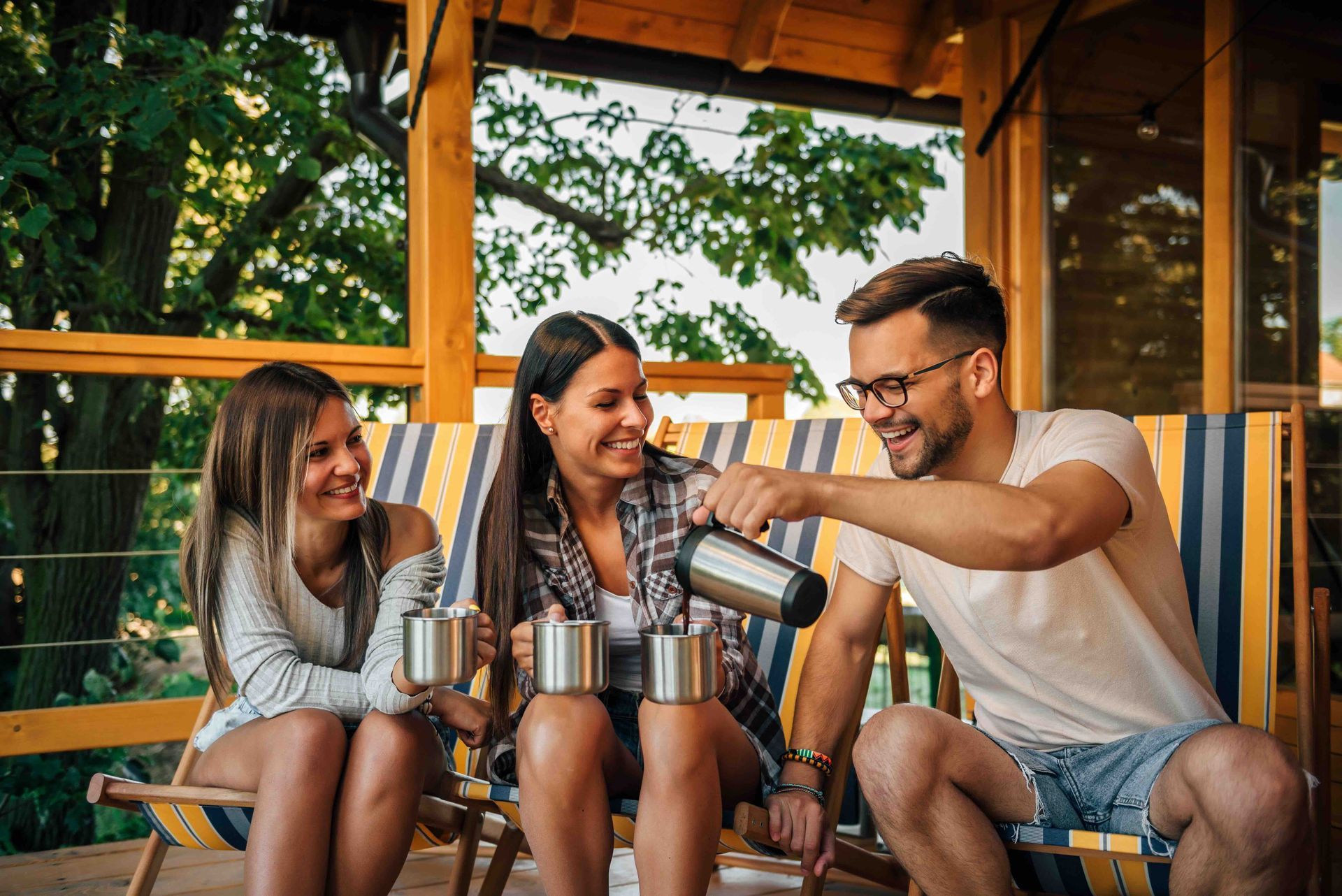 Three people on a porch, one pouring from a thermos into mugs, all smiling.