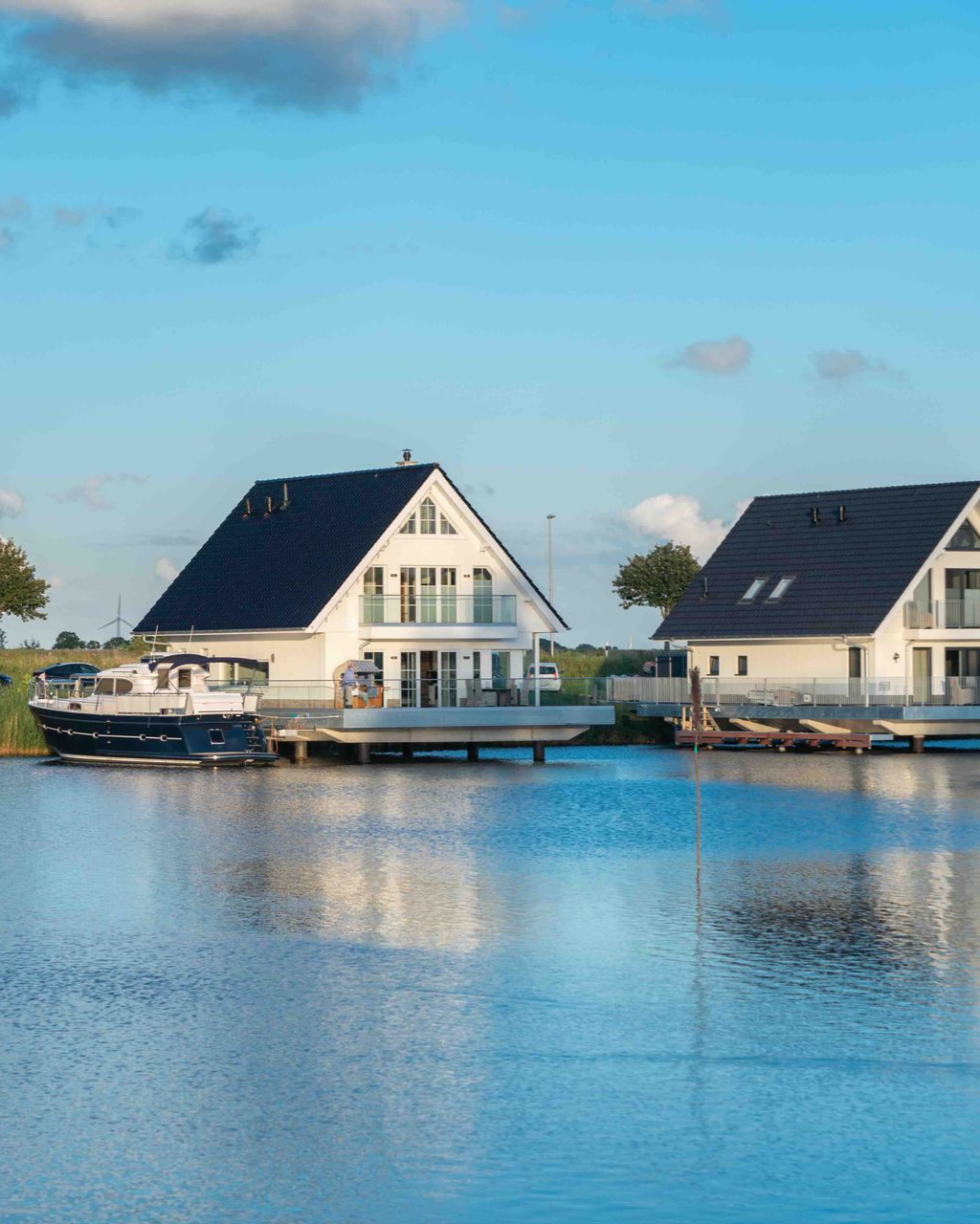 Two white houses with black roofs on a body of water, boats docked, blue sky.