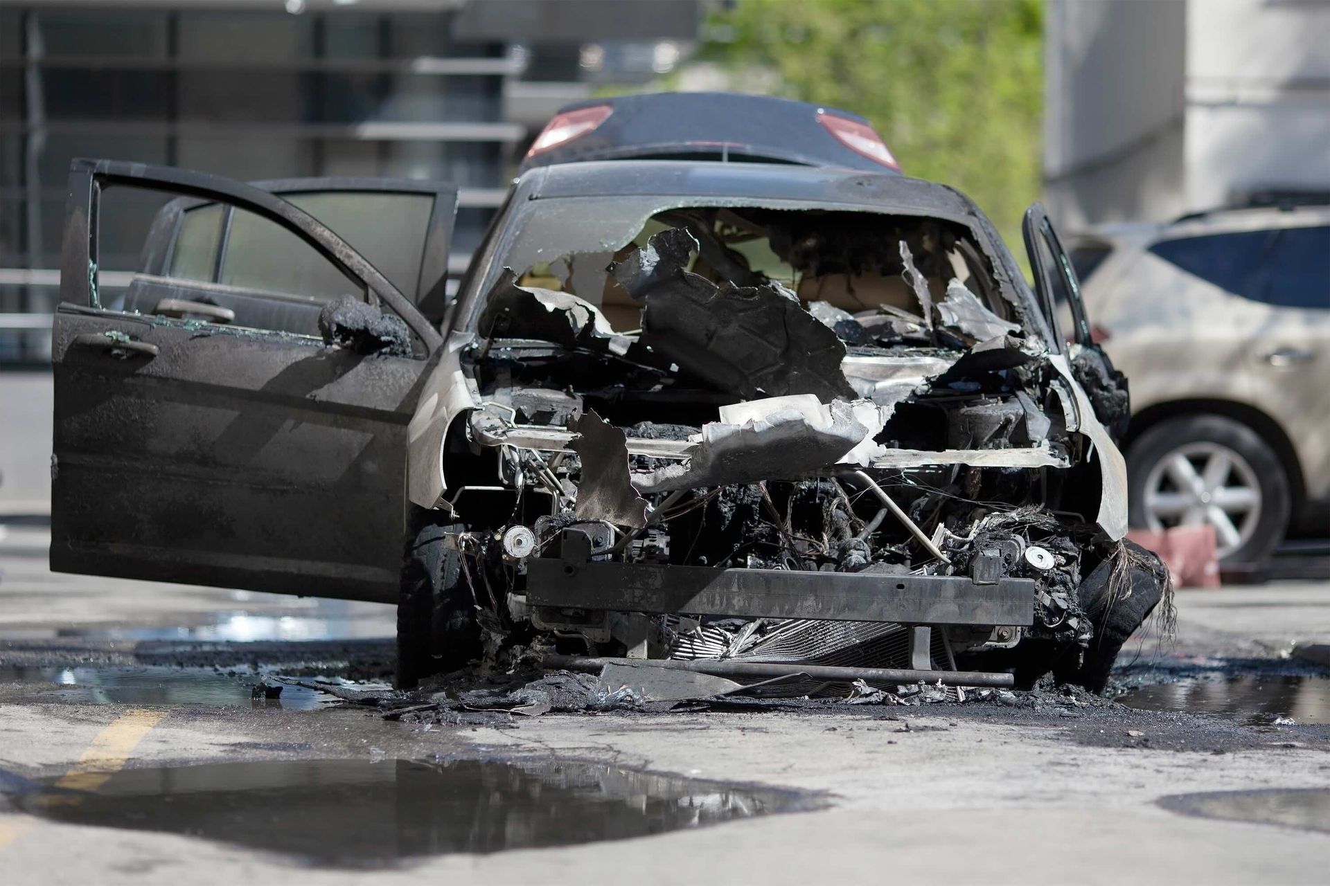Burnt-out black car with open doors, in a parking lot. Smoke and debris are visible, near another vehicle.