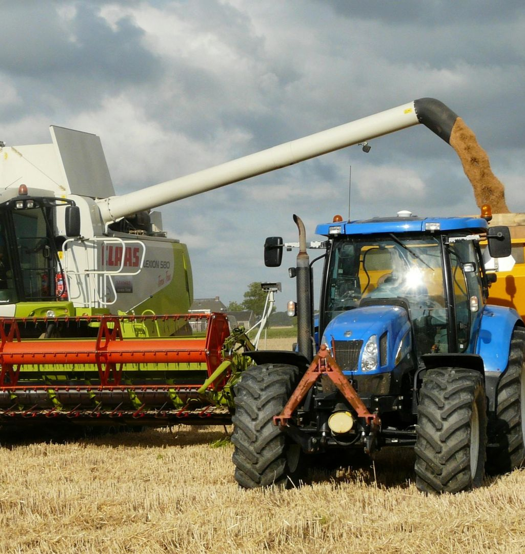 Combine harvester filling a blue tractor-trailer with grain in a field under a cloudy sky.