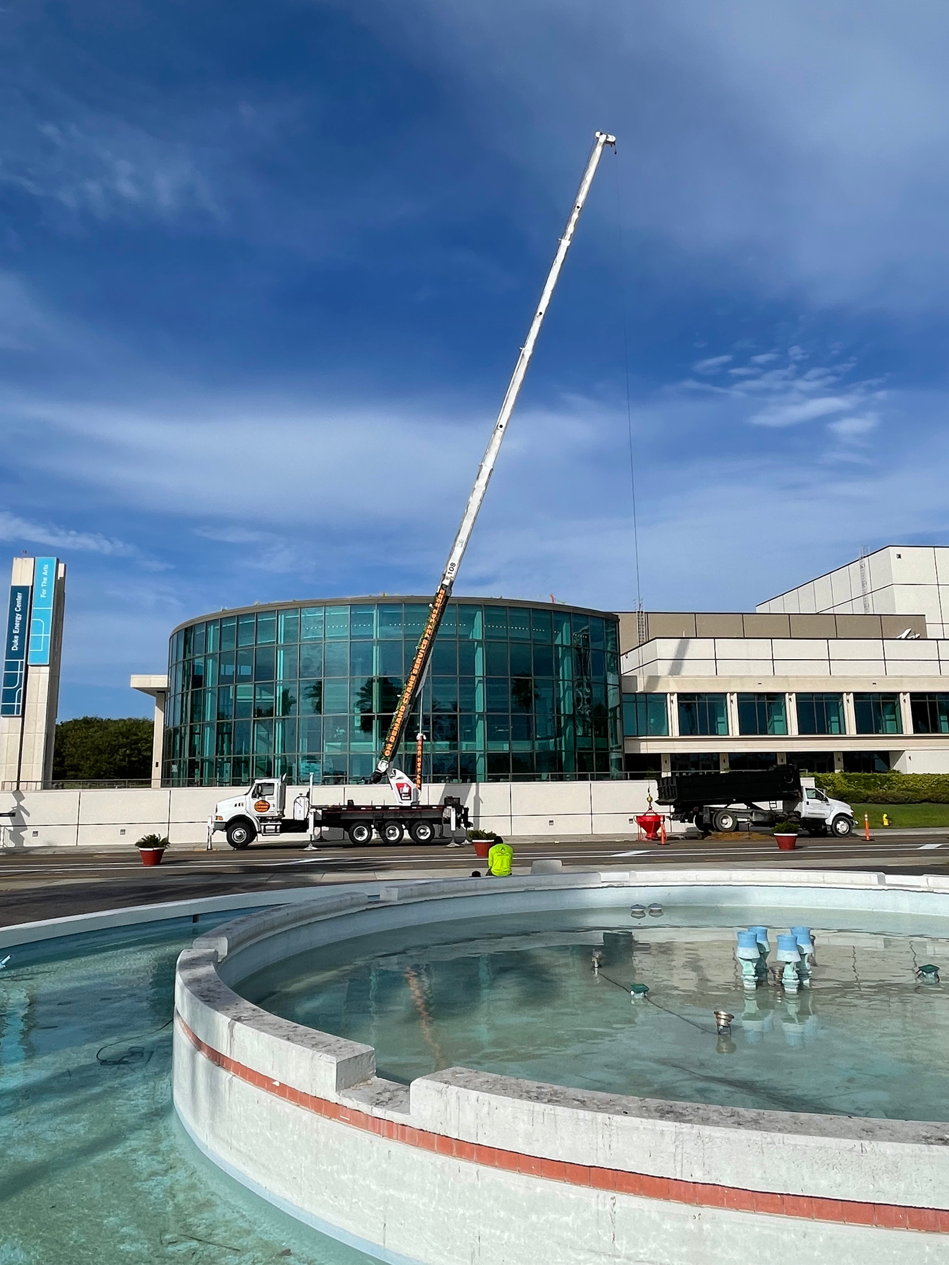 Construction crane beside a curved glass building and fountain on a sunny day