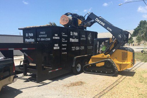 Yellow skid steer lifting wood into a black dumpster in a sunny outdoor work site