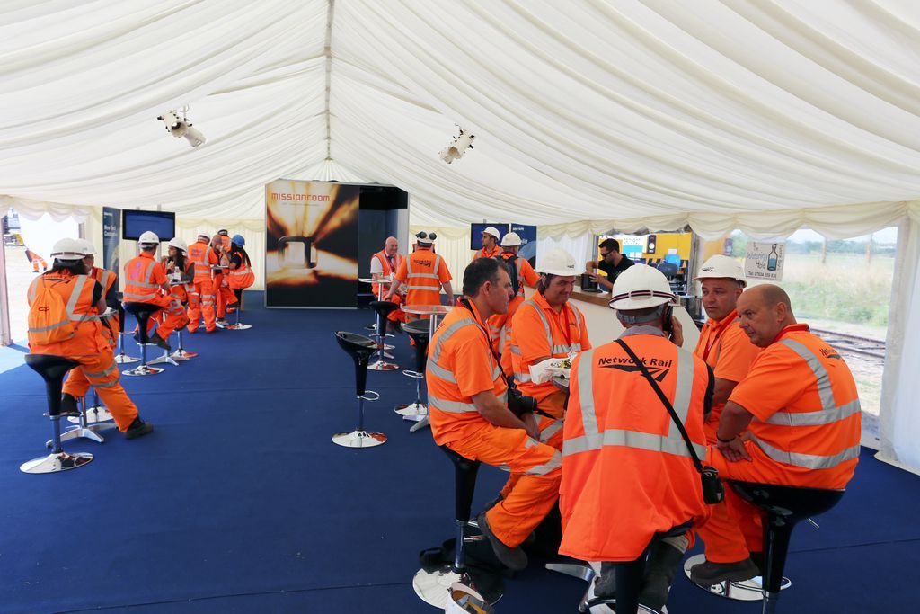 A group of construction workers are sitting at tables under a marquee