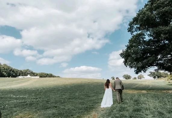 A bride and groom are walking through a grassy field.