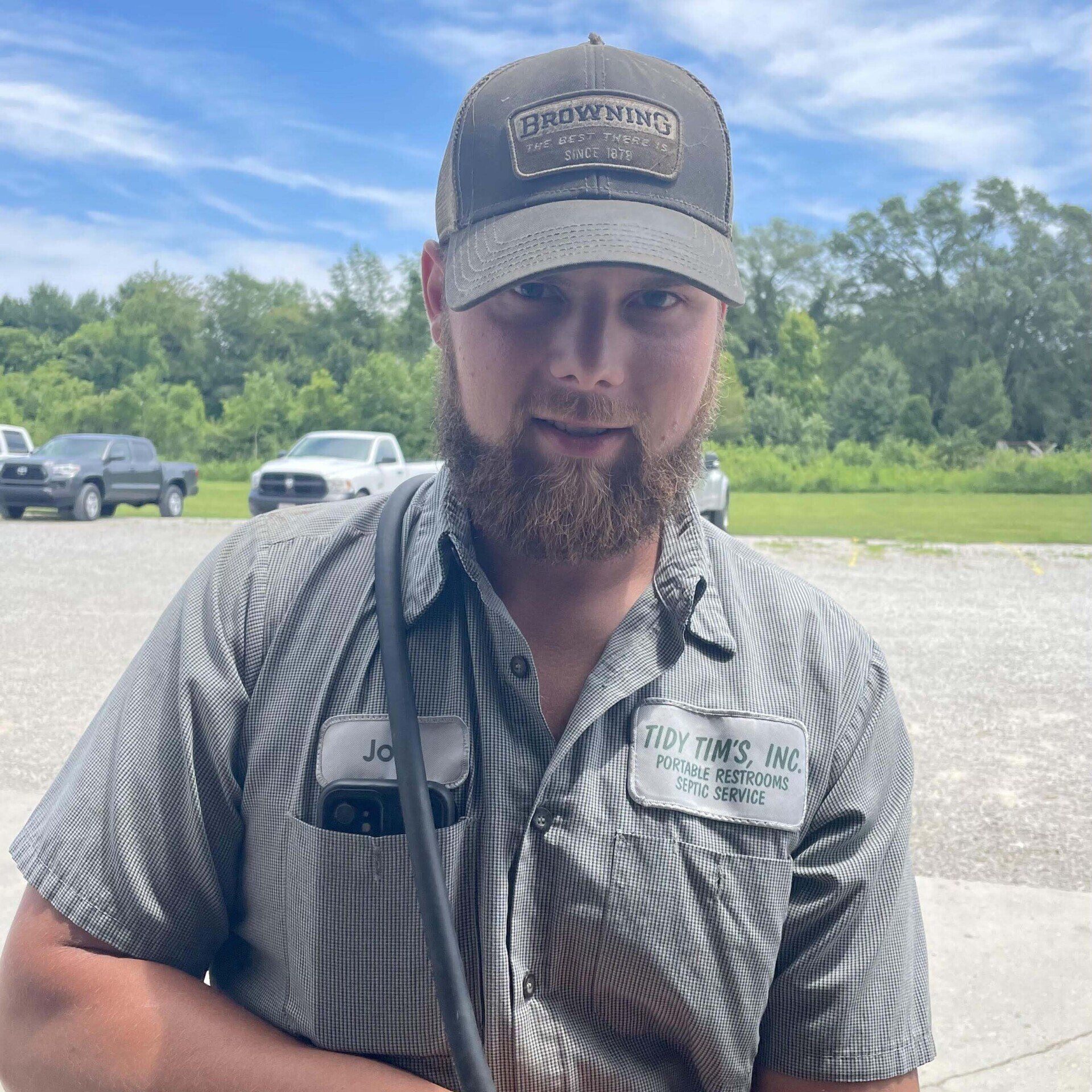 Man in cap and work shirt standing outdoors with a truck and trees in the background