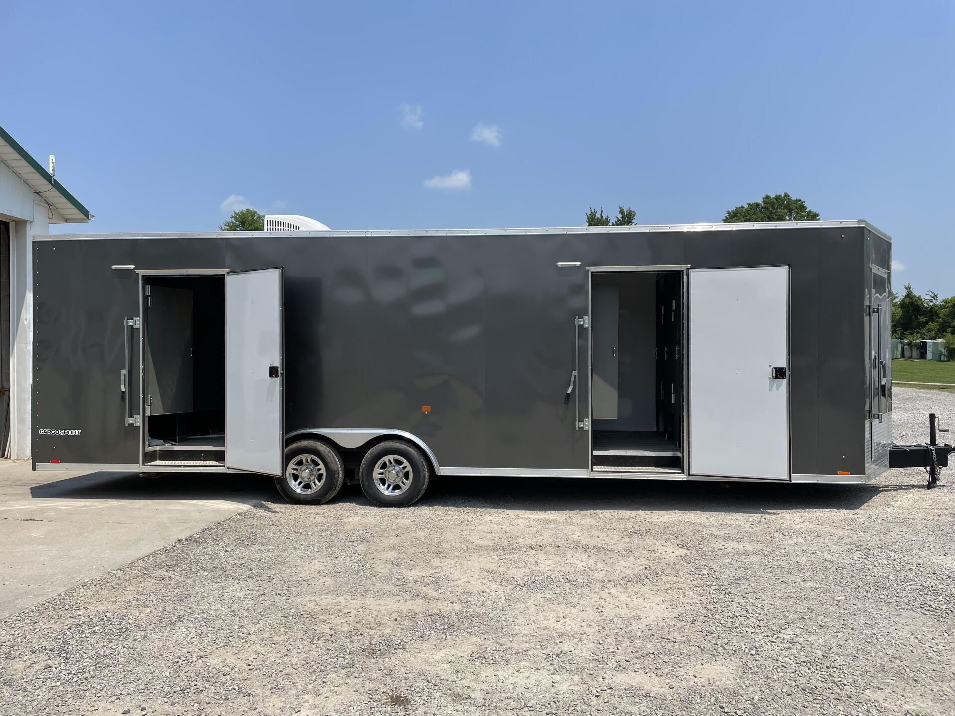 Gray enclosed trailer with two open side doors parked on a gravel lot under a blue sky