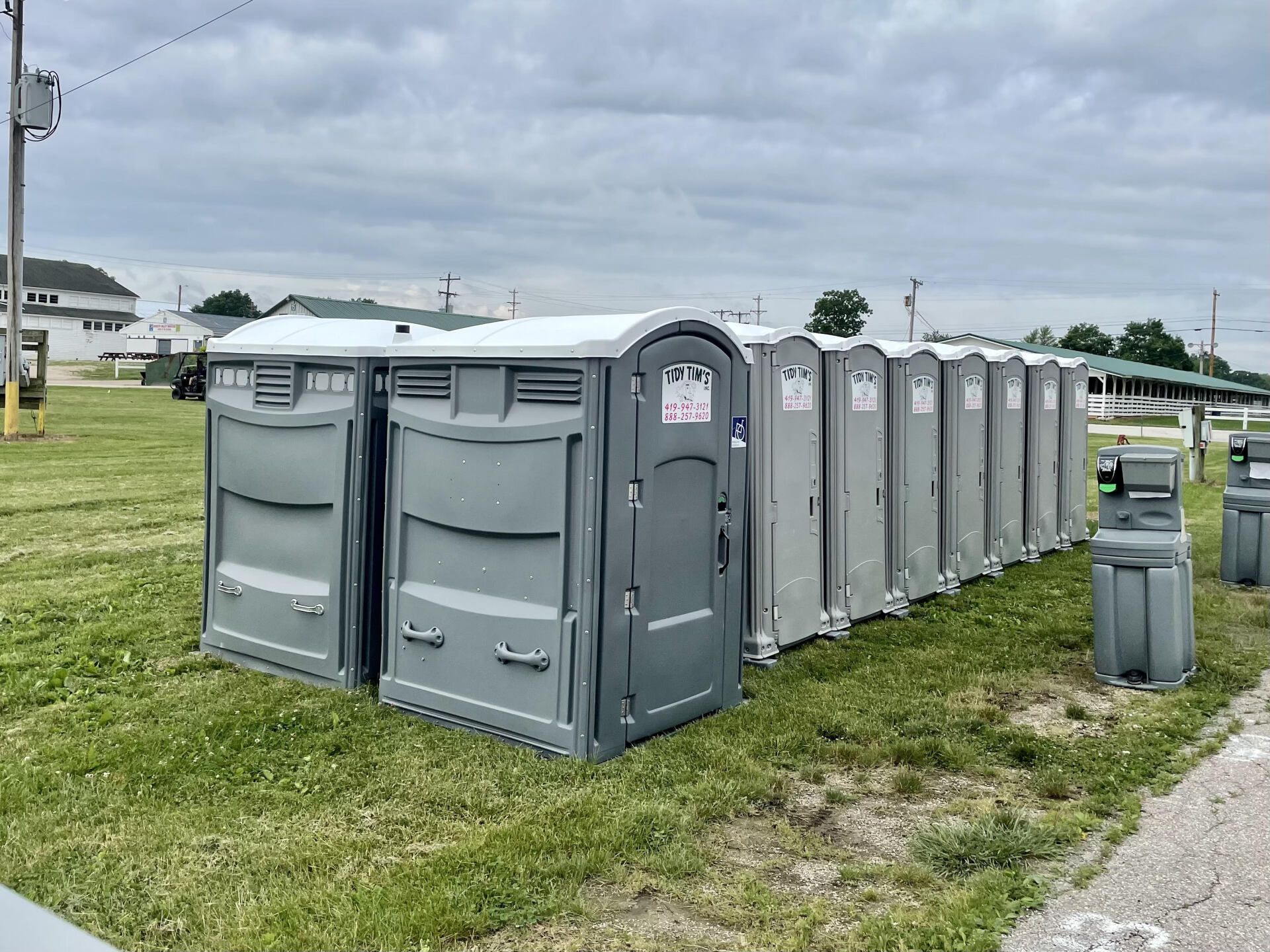 Row of gray portable toilets on a grassy roadside under a cloudy sky