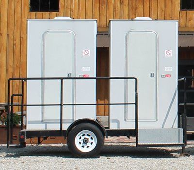 Two gray portable toilets on a black utility trailer with a white wheel, parked in front of wooden siding.
