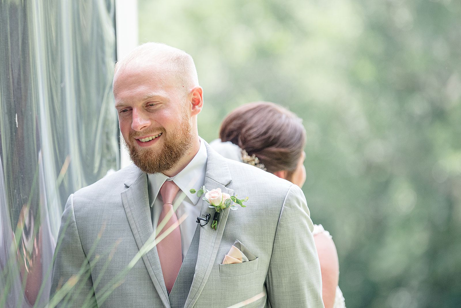 Smiling man in light gray suit and pink tie, standing outdoors with a blurred person behind him.