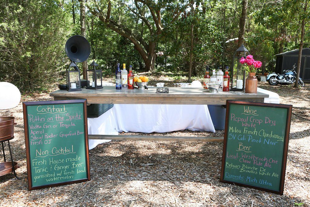 Outdoor buffet table with white cloth, chalkboard menus, and a large speaker in a wooded setting