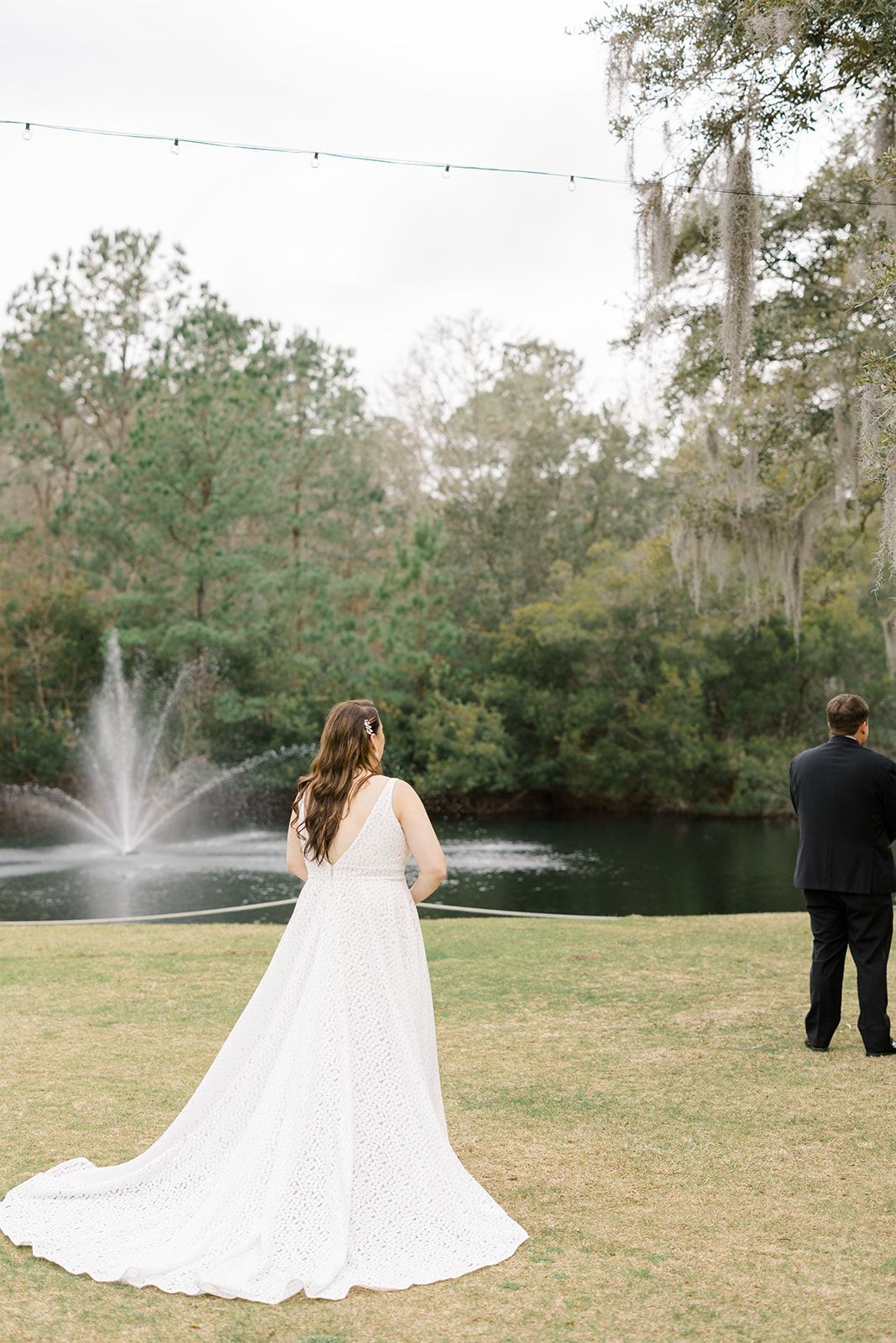Bride in a white gown and groom in a black suit stand by a pond with a fountain at an outdoor wedding.