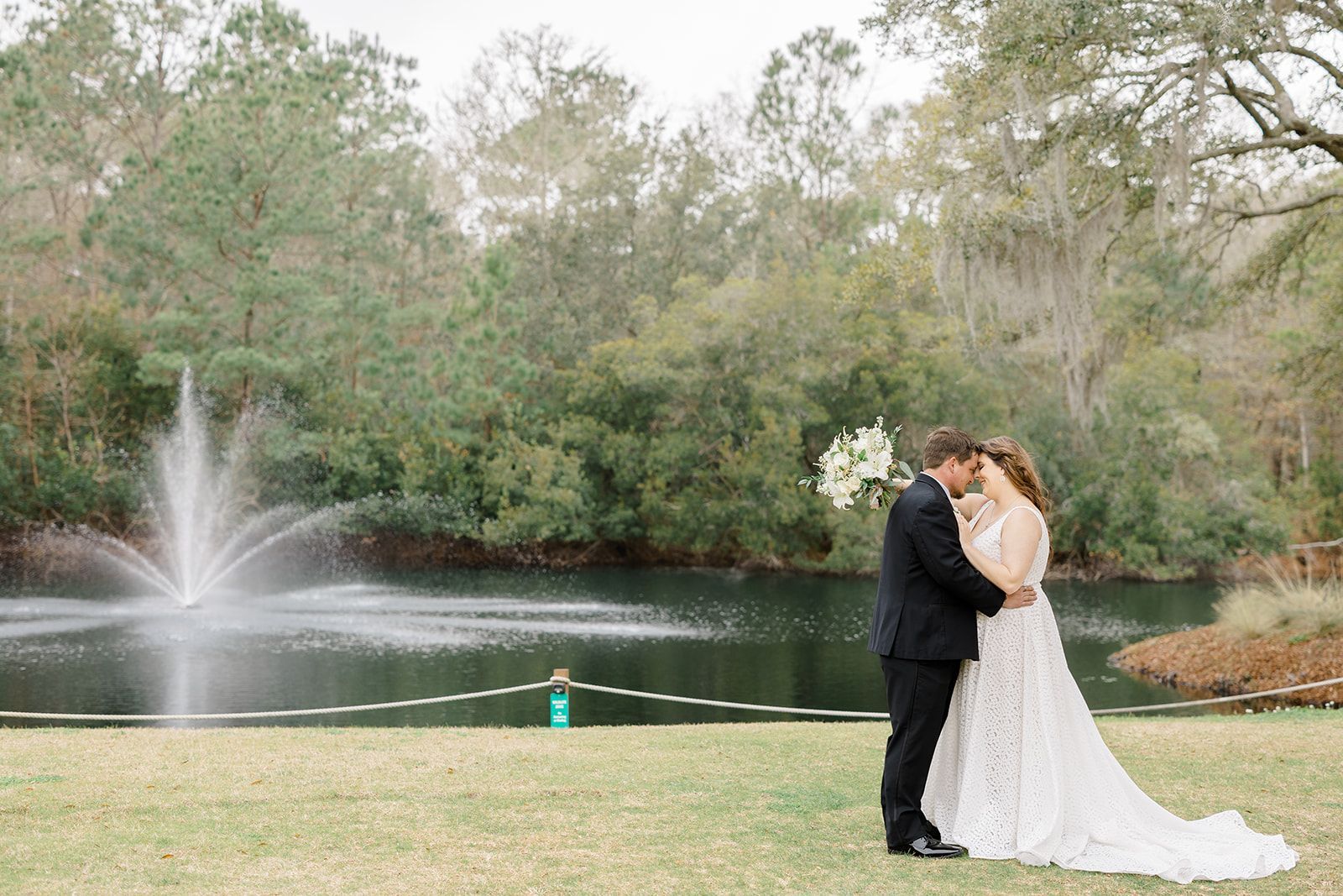 Couple in formal attire embracing by a lake with a fountain and trees in the background