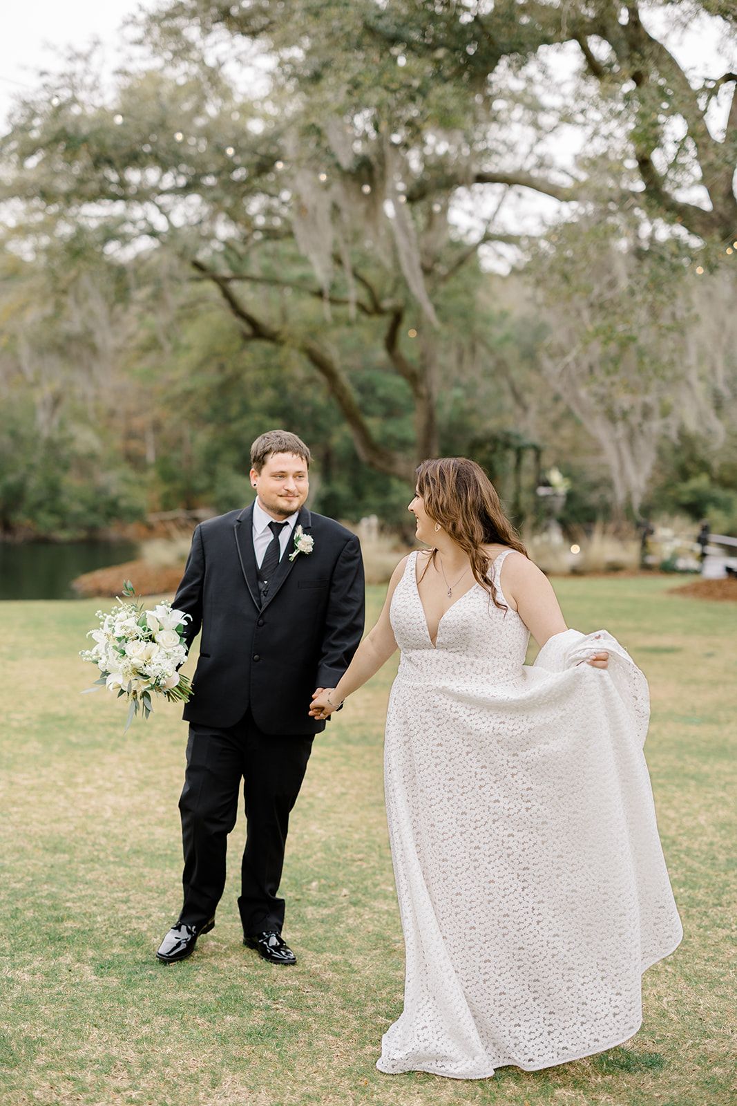 Bride and groom standing on grass outdoors, holding hands near a lake and trees.