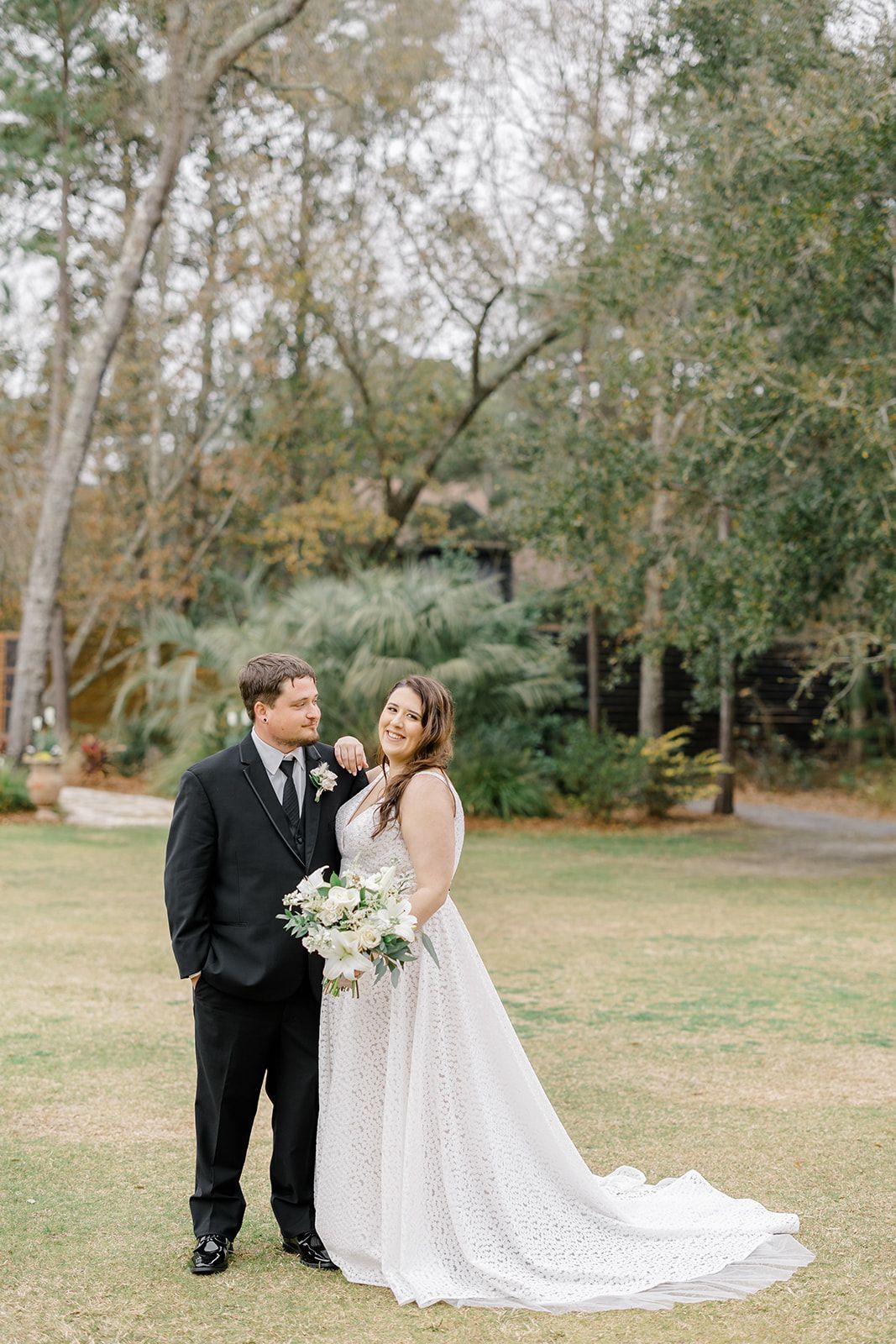 Bride and groom posing outdoors in a garden, holding a white bouquet and smiling.