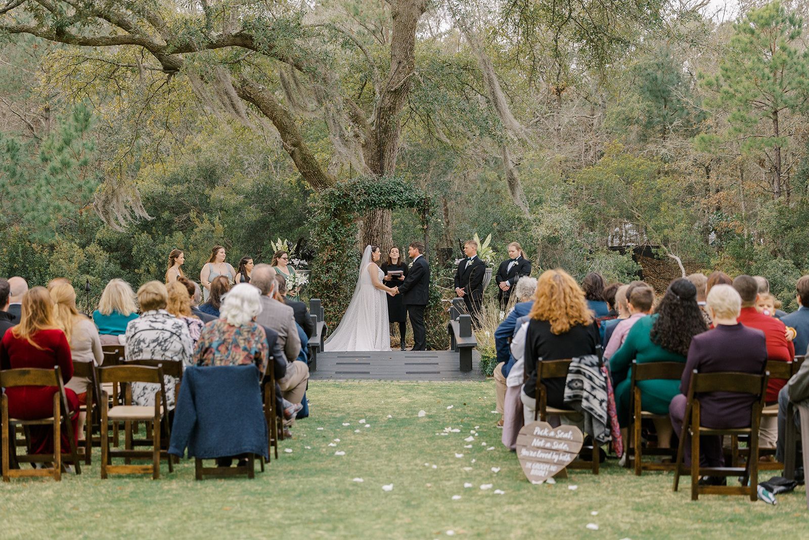 Outdoor wedding ceremony in a garden, with guests seated on chairs facing the bride and groom under trees.