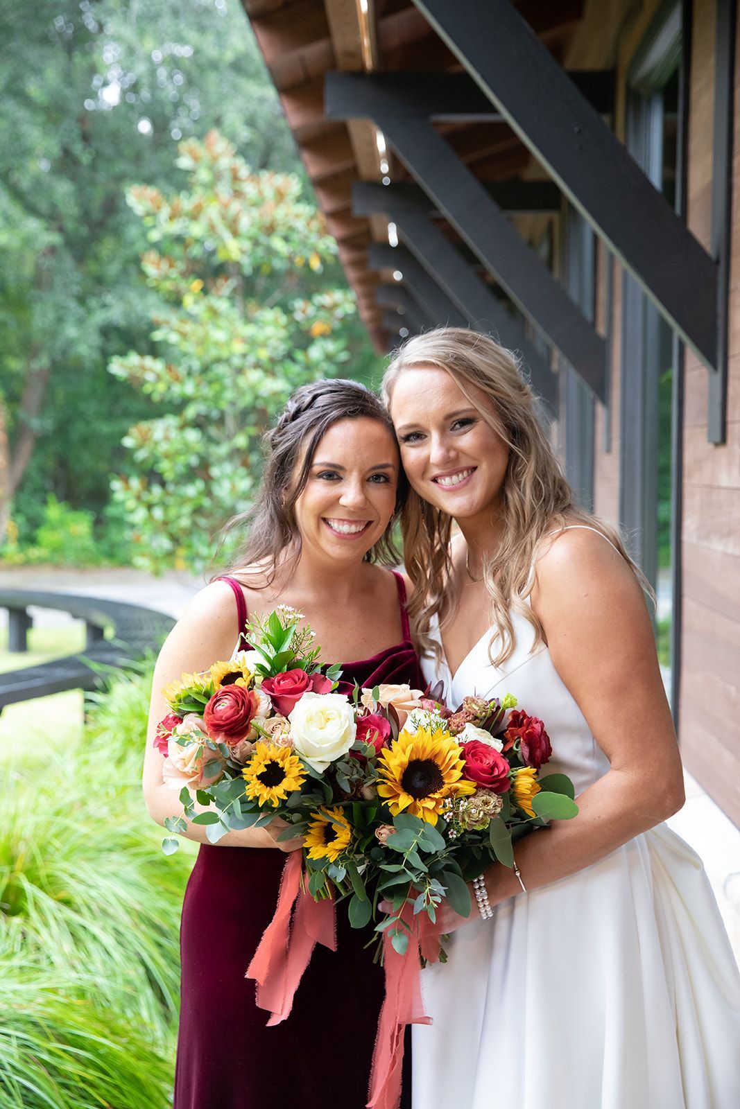 Two women in formal dresses smiling and holding colorful bouquets outdoors by a building.