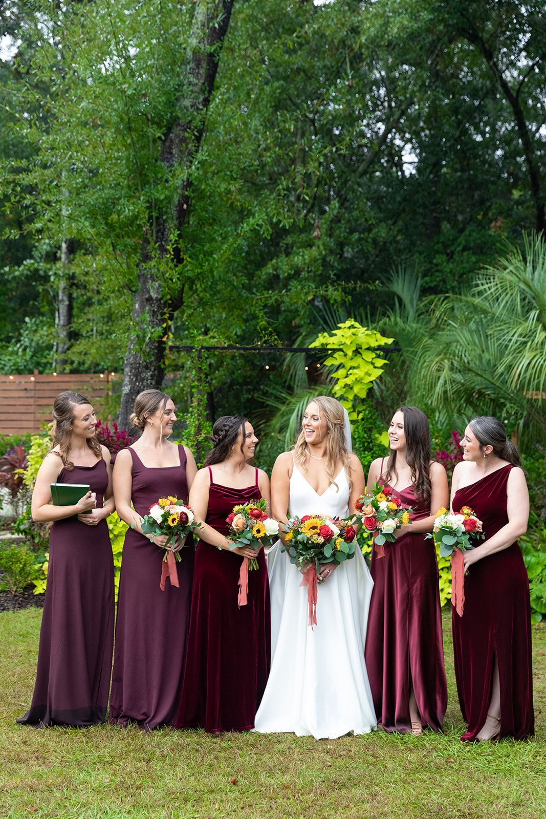 Bridal party holding bouquets outdoors, with the bride in white centered among women in burgundy dresses