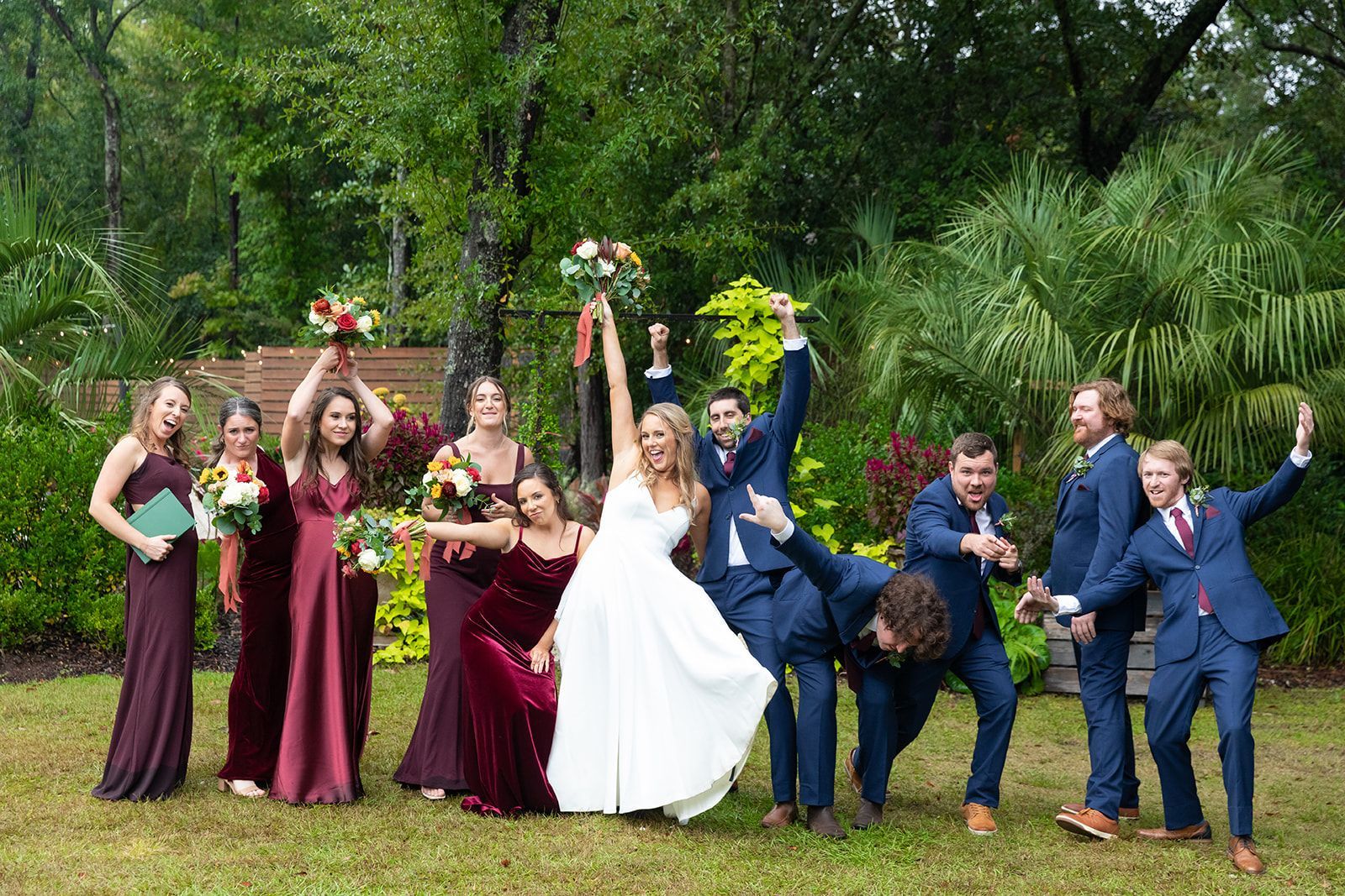 Wedding party posing outdoors on grass, with the bride and bridesmaids in burgundy and groomsmen in blue suits