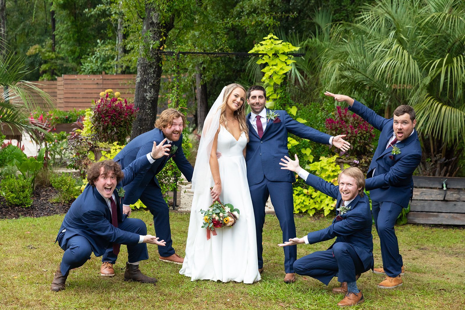 Wedding party posing playfully in a garden, with bride and groom at center in blue suits and white dress.