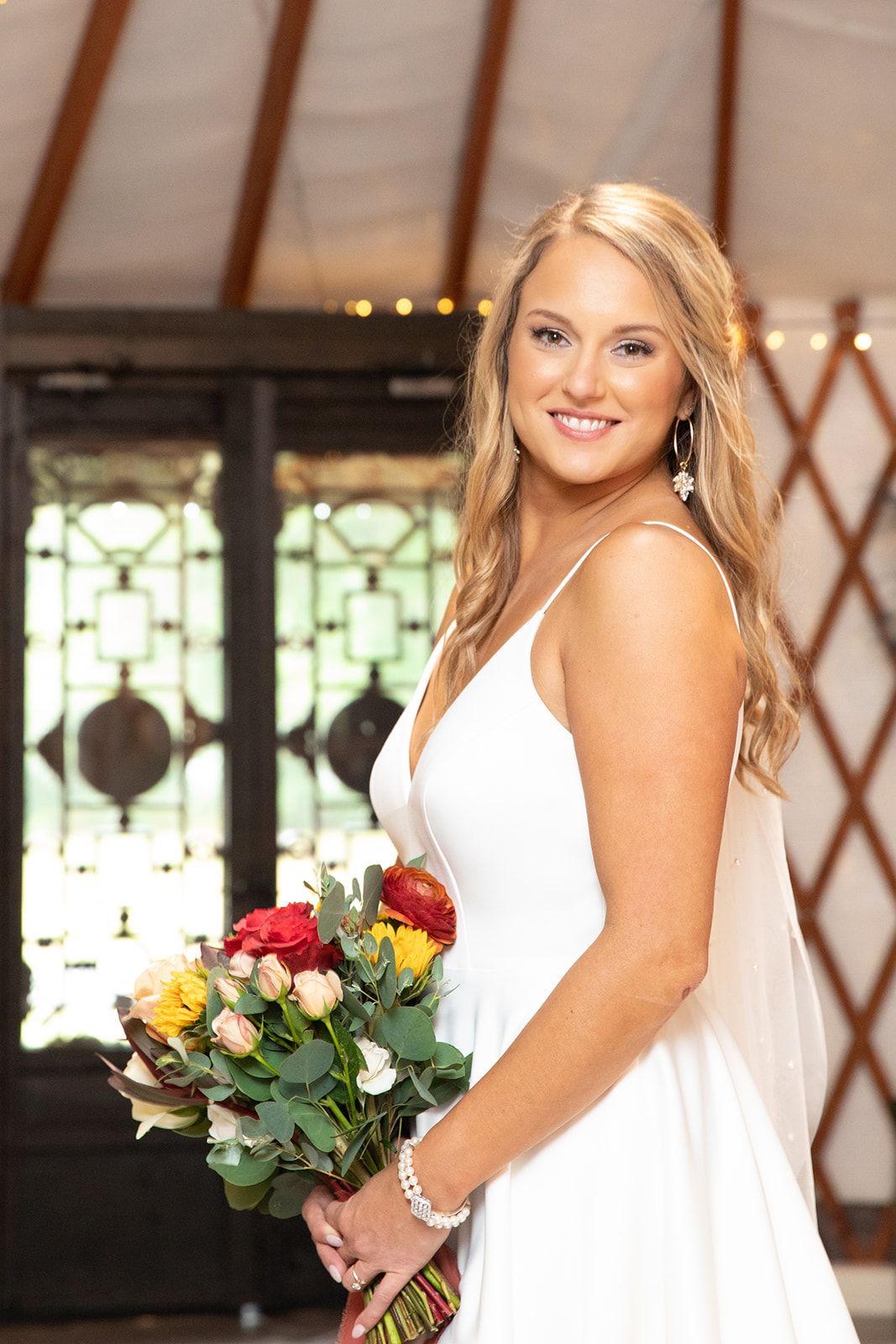 Smiling bride in a white dress holding a bouquet of red and white flowers indoors