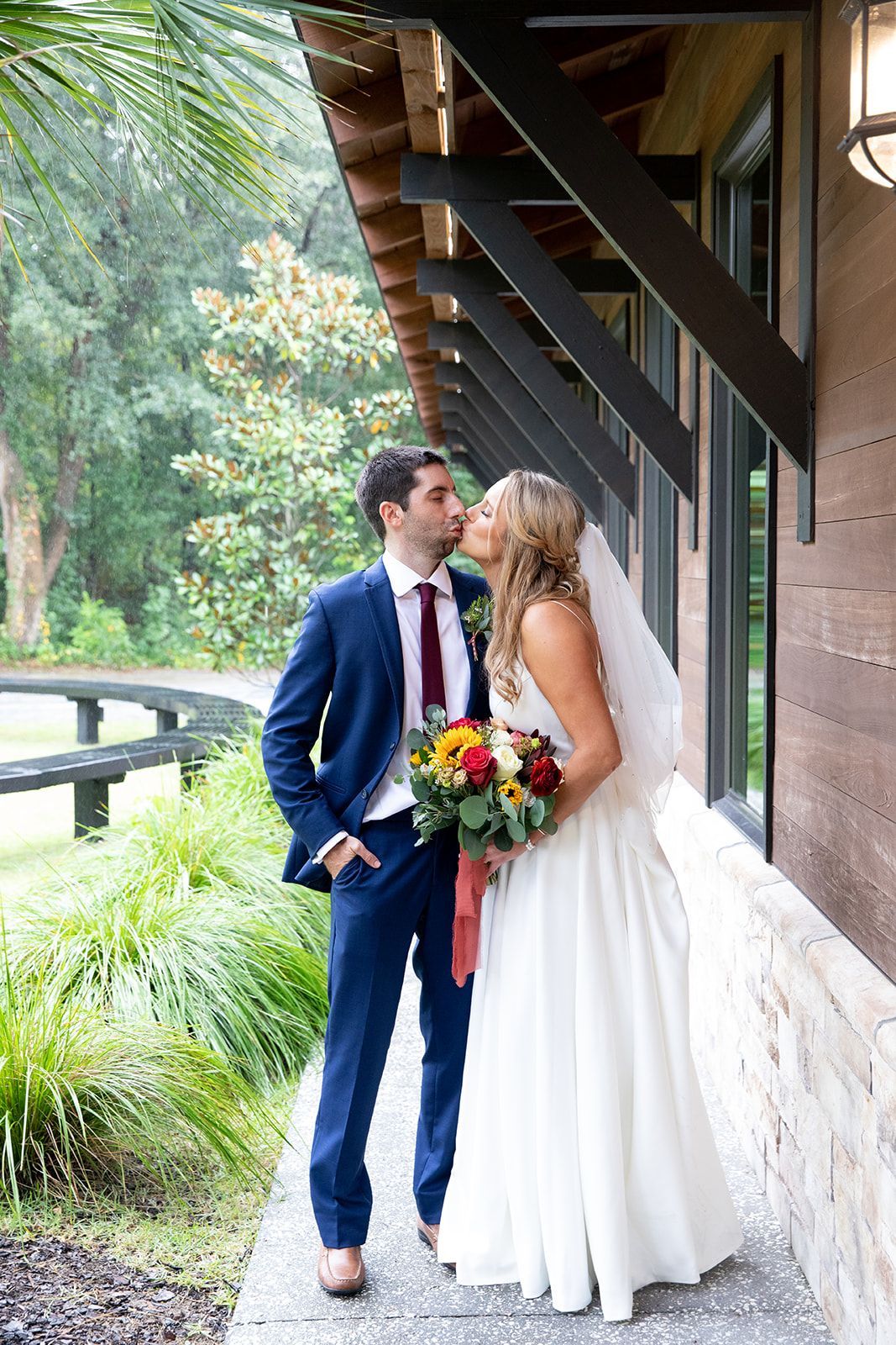 Bride and groom kissing beside a rustic building, bride holding a colorful bouquet