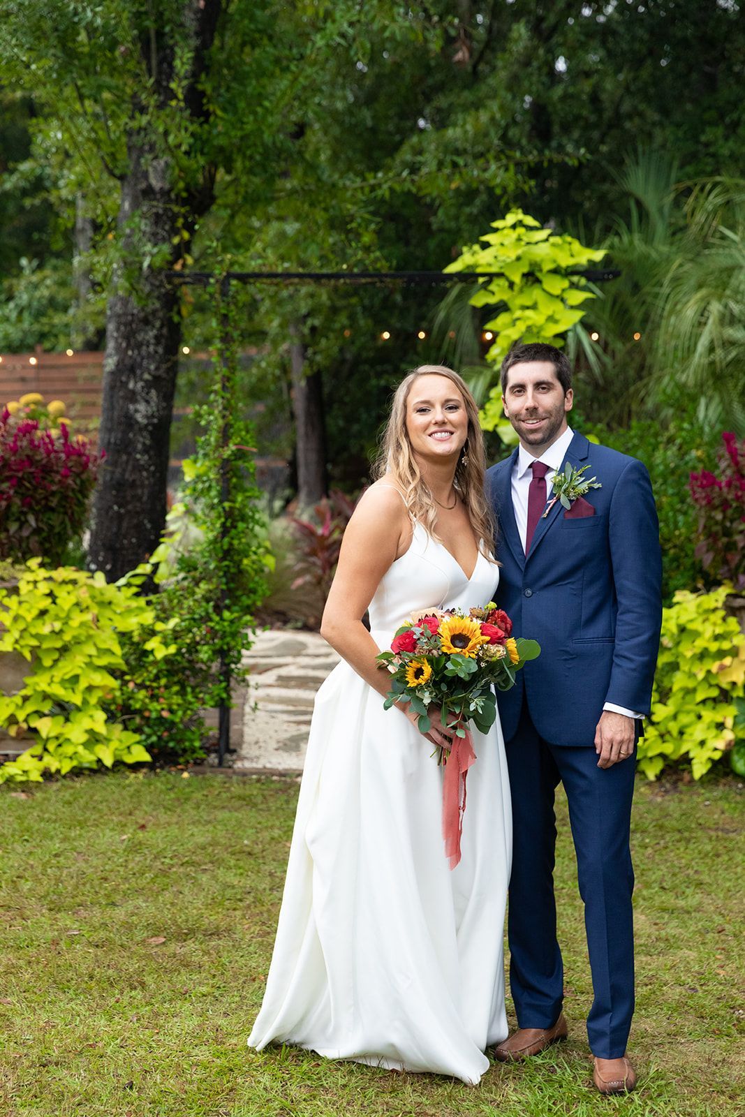 Bride and groom smiling in a garden, holding a colorful bouquet, with lush greenery behind them