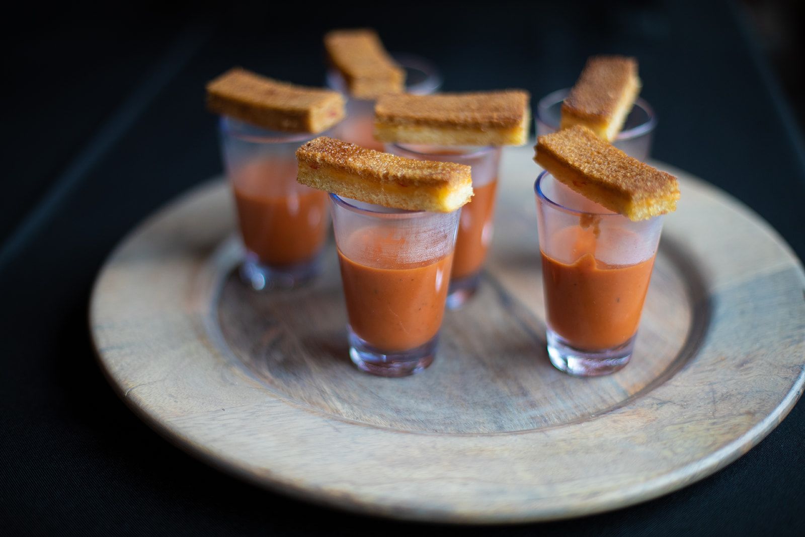Four small cocktail shots on a round tray, each topped with a toasted garnish