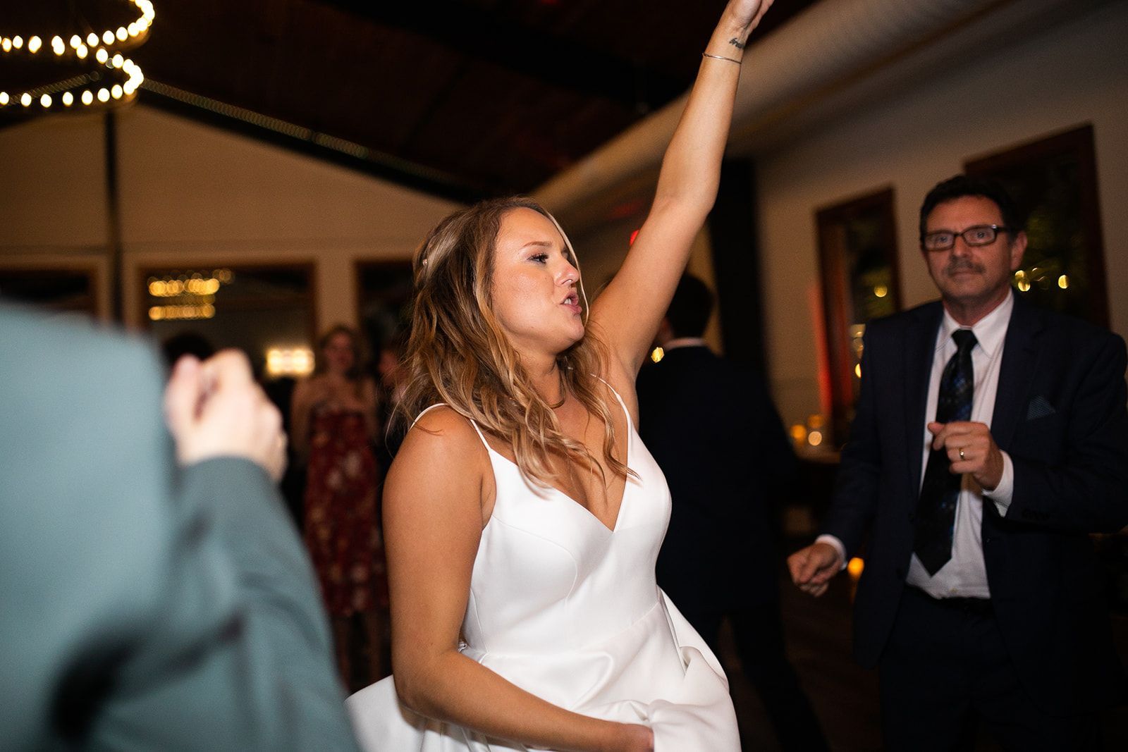 Woman in a white dress dancing with one arm raised at a dimly lit party, man in a suit nearby