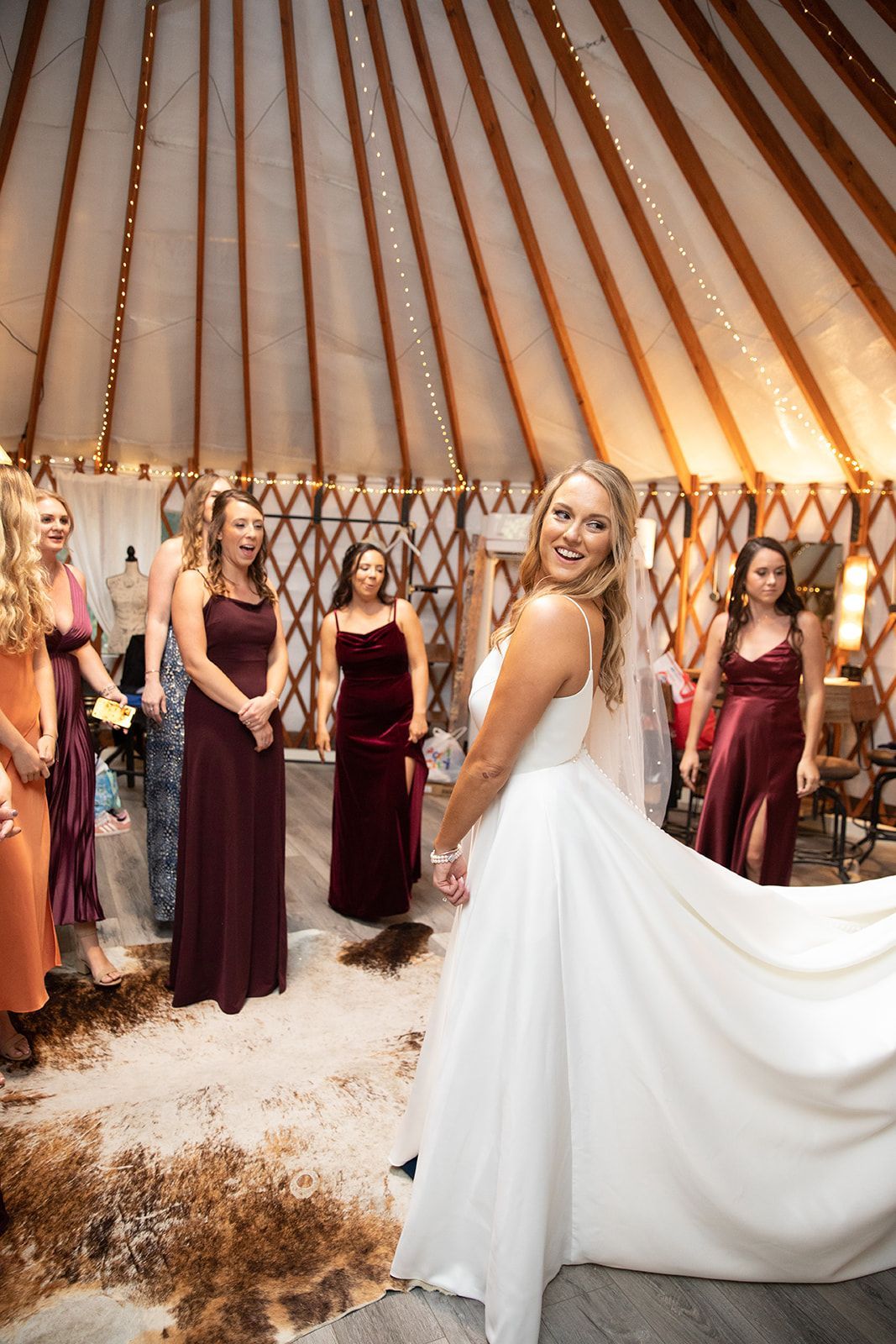 Bride in white gown turns in a tented venue as bridesmaids in burgundy dresses stand behind her.