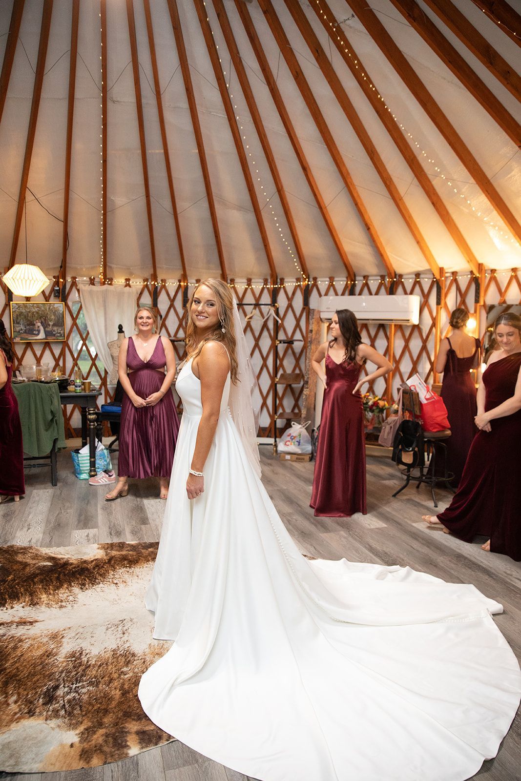 Bride in white gown with long train stands in a tent aisle beside bridesmaids in burgundy dresses
